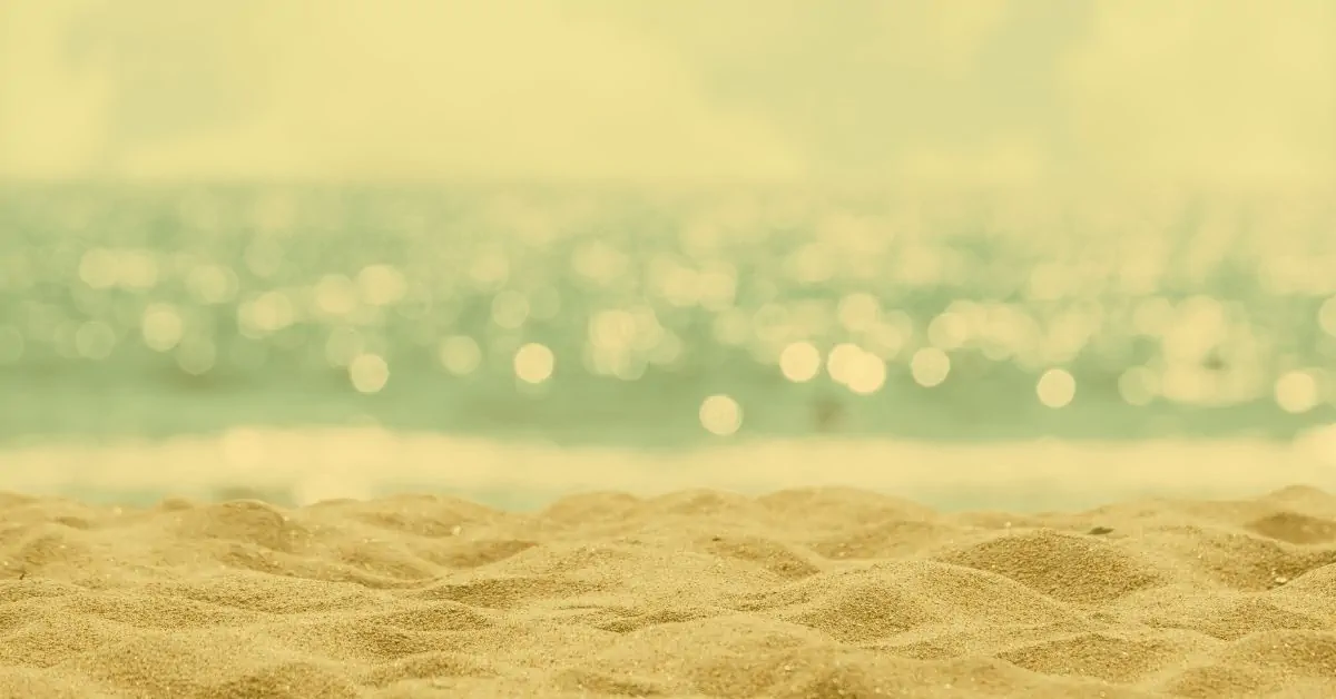 Warm golden sandy beach in the foreground leading to a softly blurred turquoise sea with gentle sunlit bokeh sparkles and pale summer sky