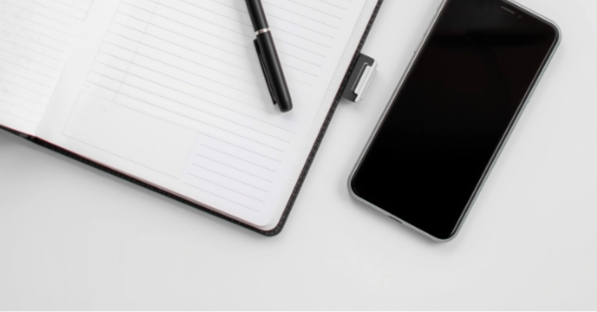 Open lined notebook with black pen resting on top beside a sleek smartphone on a clean white desk, ready for contact and notes.